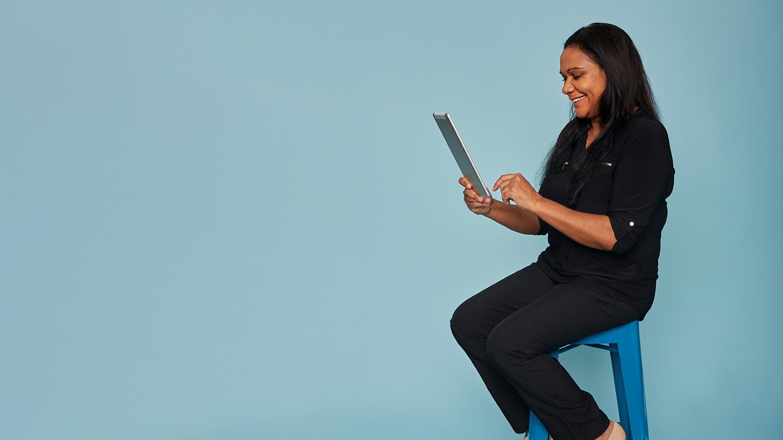 Woman sitting on a stool using a tablet, looking happy.